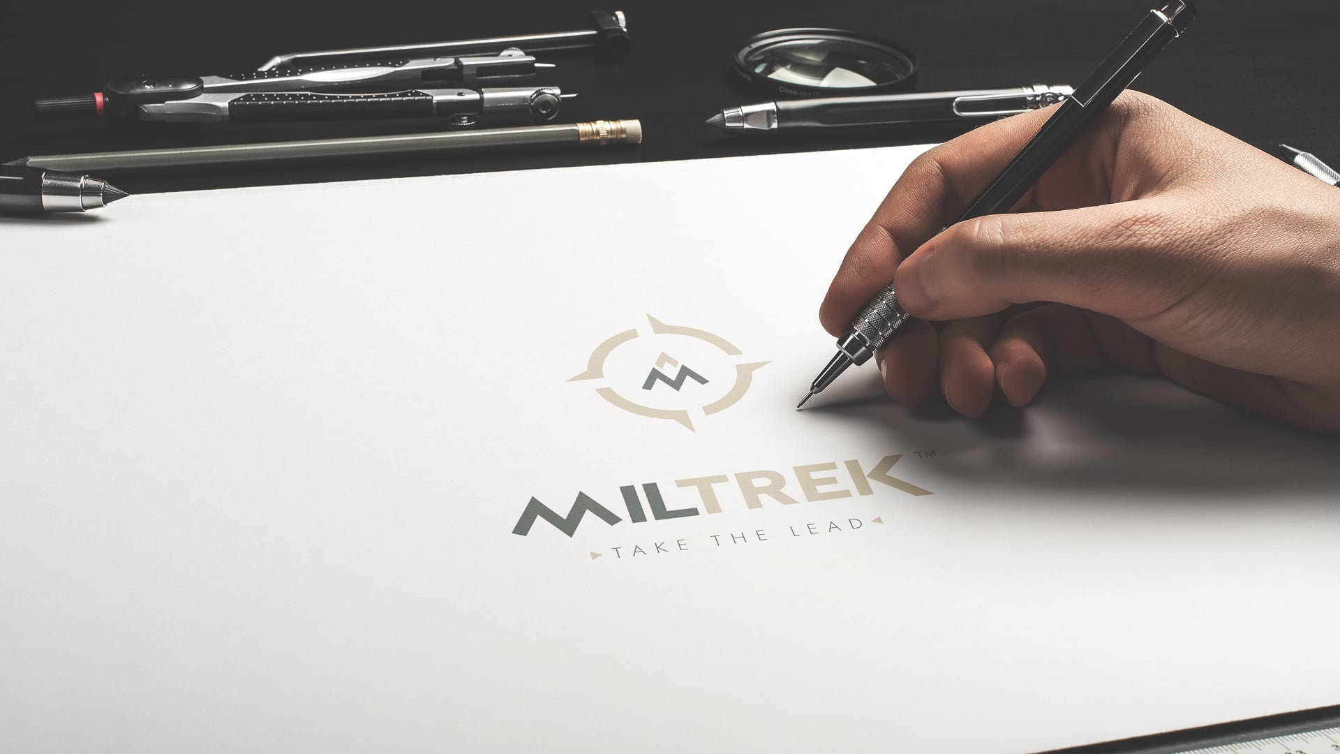 Représentation du logotype tertiaire de Miltrek sur une feuille de papier avec la main du dessinateur à côté tenant un crayon suite à la création d'identité visuelle.