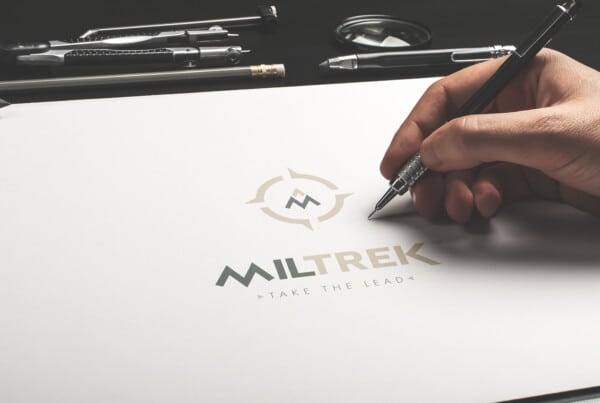 Représentation du logotype tertiaire de Miltrek sur une feuille de papier avec la main du dessinateur à côté tenant un crayon.